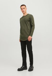 Olijfgroene blouse met lange mouwen en een ronde hals, gecombineerd met zwarte slim-fit jeans en zwarte veterschoenen, tegen een lichte achtergrond.