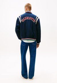 Giacca bomber blu con dettagli a strisce bianche e rosse, ricamo "Tommy Jeans" sulla schiena, abbinata a jeans blu e sneakers bianche.
