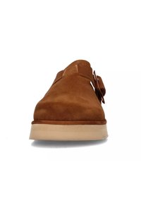 Scarpa in suede marrone con punta rotonda, texture liscia e un piccolo dettaglio d'accento. La suola è di colore chiaro e leggermente rialzata.