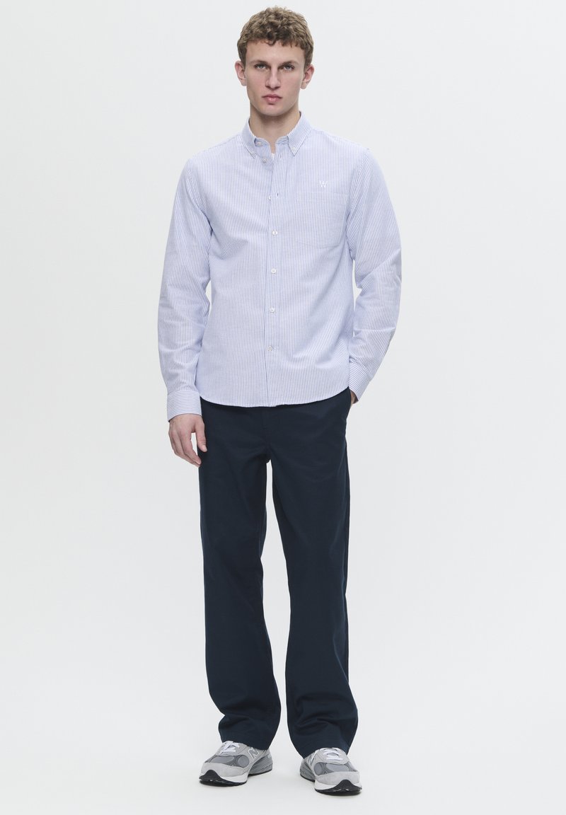 Camisa de botones a rayas azul claro con collar, combinada con pantalones oscuros de color navy y zapatillas grises. Diseño limpio, sin patrones ni acentos.