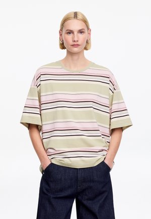 Osoba o krótkich blond włosach nosi oversize'ową pasiastą koszulkę w pastelowym różu, beżu i brązie, w połączeniu z ciemnoniebieskimi dżinsami, ręce w kieszeniach.