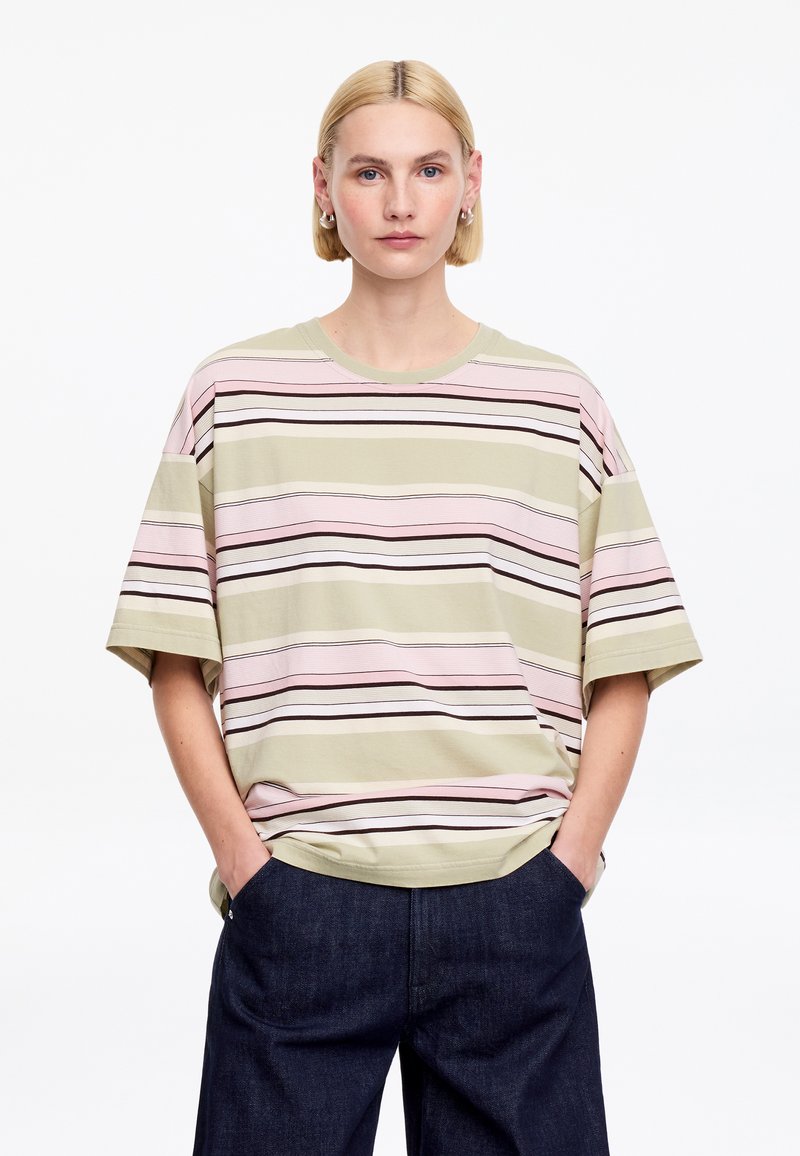 Personne aux cheveux blonds courts portant un t-shirt rayé oversize en rose pastel, beige et marron, associé à un jean bleu foncé, mains dans les poches.