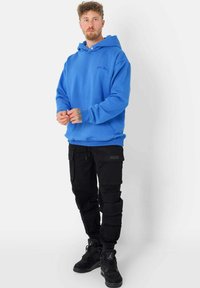 Blauwe fleece hoodie met een grote capuchon, gedragen met zwarte cargobroek met meerdere zakken en zwarte sneakers met gestructureerde accenten.