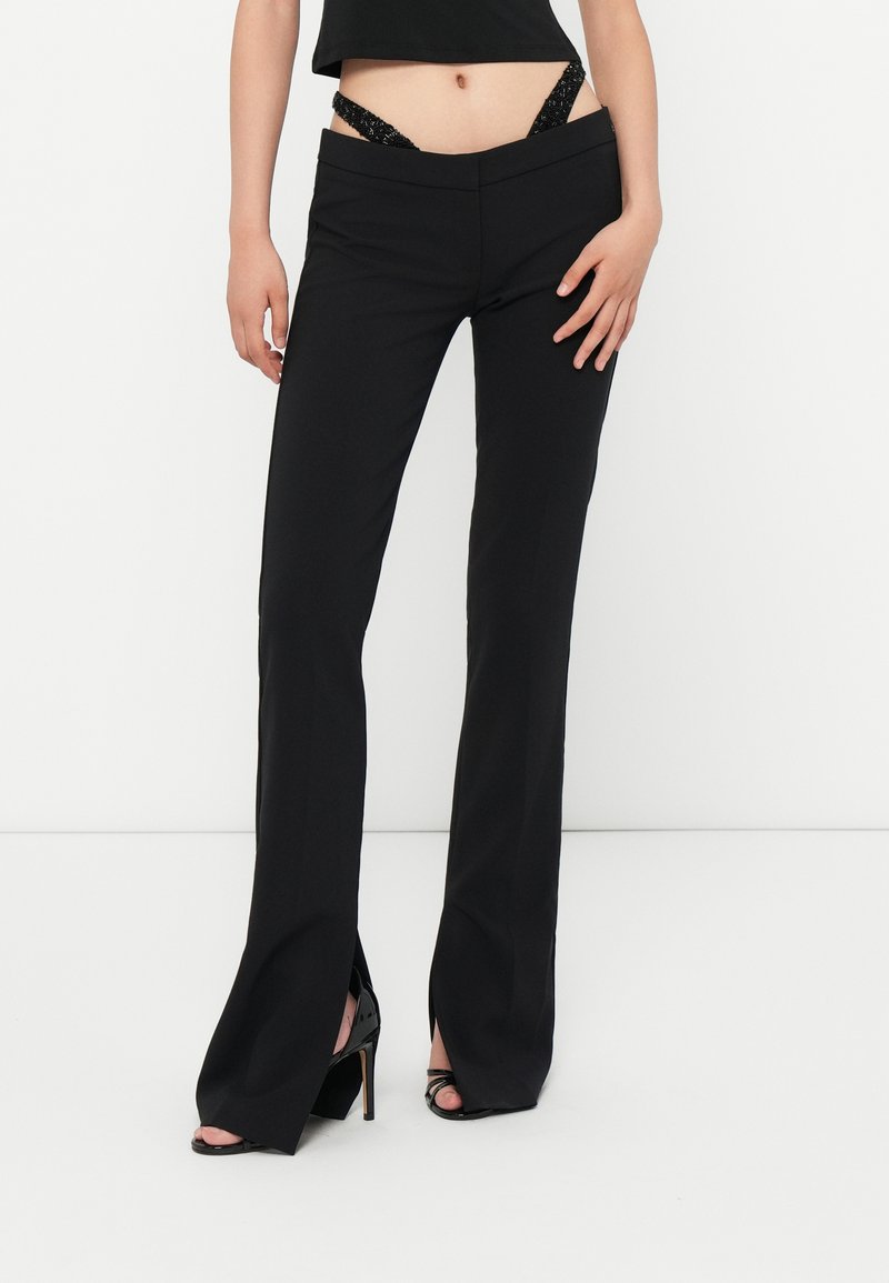 Donna che indossa pantaloni neri a vita alta con spacco, con visibile biancheria intima nera in pizzo e sandali neri con tacco alto su uno sfondo bianco.