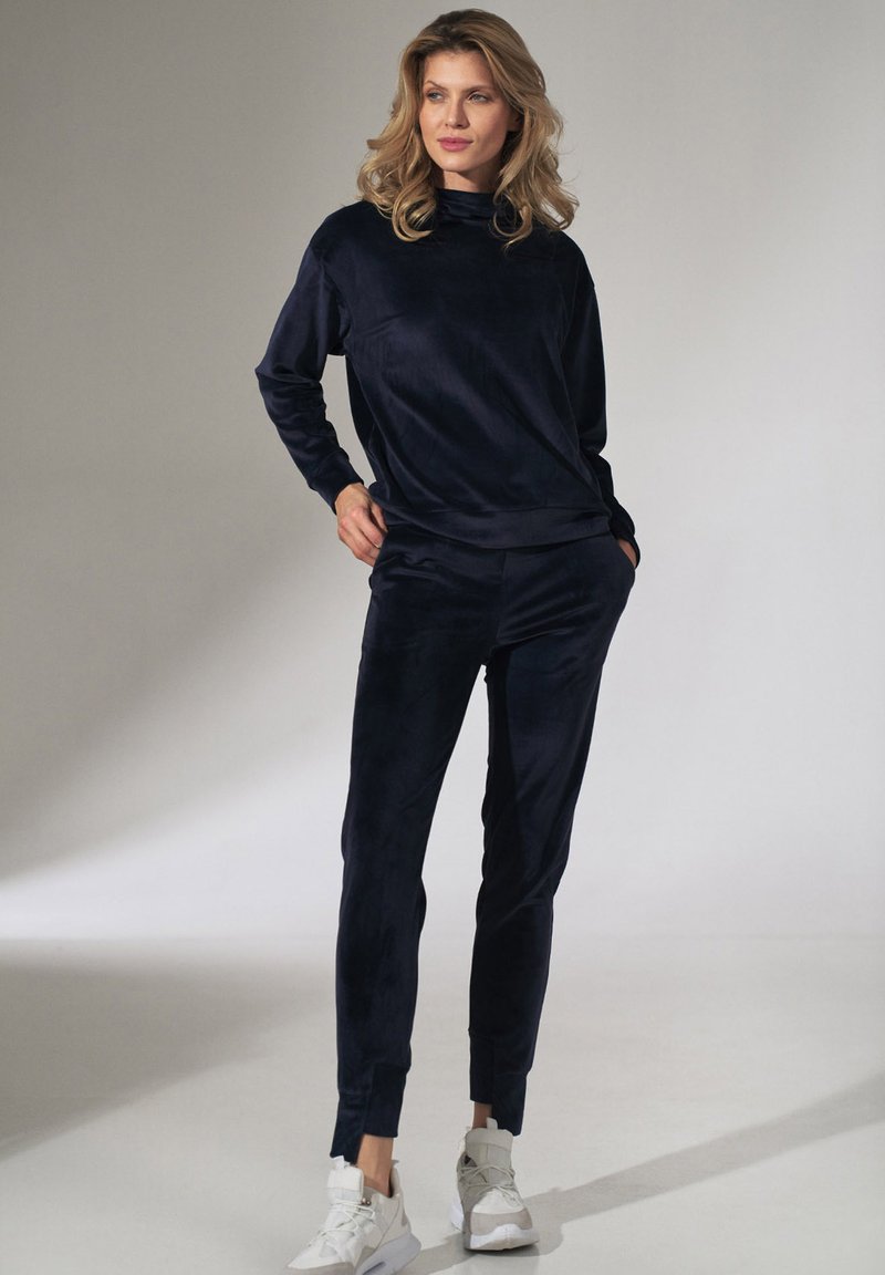 Navy blauwe velours trainingspak met een sweatshirt met hoge nek en gesneden broek, gecombineerd met witte sport schoenen. Glad textura en comfortabele fit.