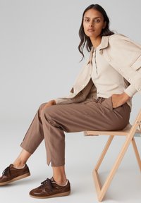 Giacca di suede beige sopra un top fitted a costine beige, abbinata a pantaloni marroni e sneakers marroni con dettagli bianchi. Texture e colori semplici.