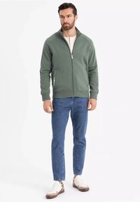 Chaqueta verde con cremallera y alto cuello, puños acanalados y bolsillos laterales, combinada con jeans azules y zapatillas en blanco con detalles en marrón.