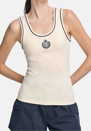 Donna che indossa una canottiera color crema con rifinitura nera e stemma ricamato, abbinata a pantaloncini cargo blu navy, mani poi riposanti sui fianchi.