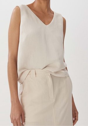 Kobieta ubrana w bezrękawnikowy, jasnobeżowy top z dekoltem w serek, wsunięty w dopasowane spodnie z wysokim stanem na gładkim tle.