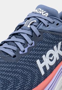 Blauer Sportschuh mit einem strukturierten Mesh-Obermaterial, HOKA-Logo und einer pinkfarbenen Verlaufssohle. Die Schnürsenkel sind flach und eng beieinander.