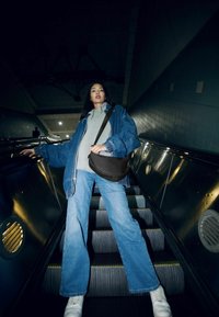 Giacca di jeans sopra una felpa grigia, abbinata a jeans azzurri e sneakers bianche. Borsa a tracolla nera, ambientazione su scala mobile con illuminazione ambientale.