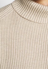 Maglione a coste beige con colletto alto, tessuto testurizzato e righe verticali. Primo piano sui dettagli del materiale e della cucitura.