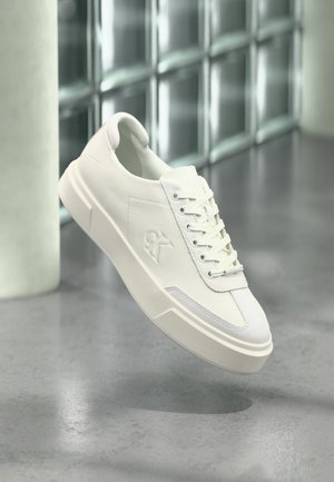 Bela nizka superga z debelim podplatom, reliefnim logotipom na strani in vezalkami, prikazana v sodobnem, odsevném notranjem okolju.