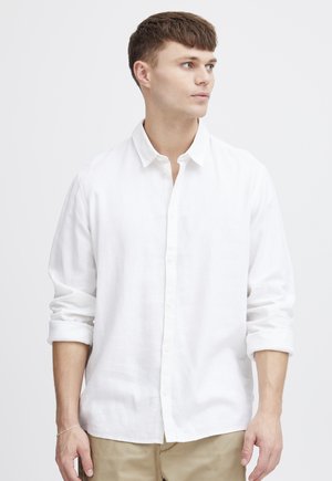 Ung mand med kort brunt hår iført en hvid skjorte med knapper og opsmøgede ærmer samt beige bukser, der kigger mod højre.