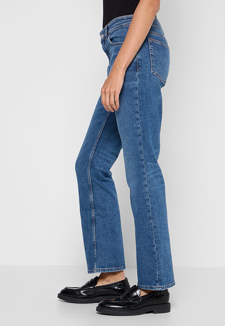 Persoon in een blauwe rechte jeans, zwarte loafers en een zwart shirt, zijwaarts staand tegen een plain witte achtergrond.