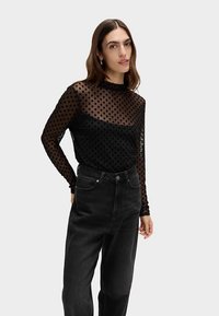 Black long-sleeve top with sheer polka dot fabric, layered design, and a smooth black underlayer. Worn with high-waisted black jeans.