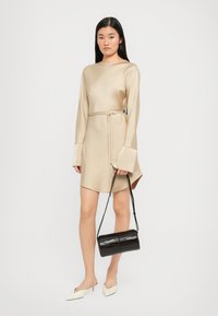 Beige pitkähihaisen mekko, jossa on vyötäröllä vyö ja liehuvat hihansuut, yhdistetty valkoisiin kärkitossuihin ja mustaan krokotiilikuosisaksi.