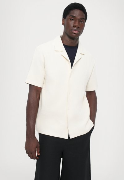 Kurzärmliges, knöpfbares Hemd aus strukturiertem cremfarbenem Stoff. Hat einen Kragen und einen lockeren Schnitt, kombiniert mit schwarzen weit geschnittenen Hosen.