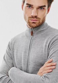 Szary sweter na zamek z wysokim kołnierzem, ściągaczami na rękawach i brązowym skórzanym uchwytem zamka; materiał wygląda na miękki i teksturowany.