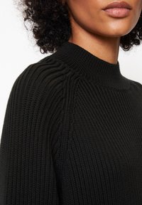 Pull noir en maille avec un col montant côtelé, présentant des rayures verticales texturées et une silhouette ajustée. Aucun matériel visible ni ornements.