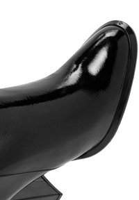 Bottines en cuir verni noir avec un bout pointu et un talon géométrique empilé. Texture lisse et brillante avec des détails de couture près du talon.