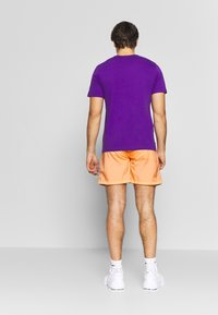 Fialové bavlněné tričko spárované s oranžovými šortkami. Model stojí zády, obutý v bílých sportovních botách a kotníkových ponožkách. Jednoduchý design, bez vzorů.