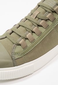 Baskets vert olive avec empeigne en maille texturée, bout en caoutchouc lisse et lacets plats. Semelle en caoutchouc blanche avec détails côtelés.