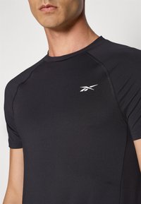 T-shirt de sport noir avec des manches courtes, un design ajusté et un petit logo blanc sur la poitrine. Texture lisse avec des coutures plates.