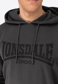 Grå hættetrøje med snøre i hætten, prydet med et stort sort "LONSDALE" logo og "LONDON" tekst foran. Blødt, tekstureret stof.