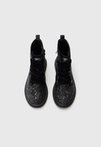 Botas negras hasta el tobillo con una superficie brillante, diseño con cordones y acentos de terciopelo, con suela de goma negra y detalle de cremallera lateral.