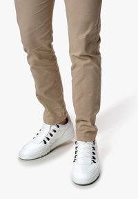 Sneaker bianche con finitura in pelle liscia, caratterizzate da occhielli e lacci neri, abbinate a pantaloni slim-fit beige chiaro.