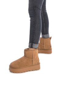 Botas de tobillo de ante marrón con un forro suave, punta redonda, detalles de costura y suela de goma texturizada, mostradas con jeans oscuros enrollados.