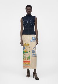 Mannequin portant un haut en denim foncé sans manches avec des formes de cœur effilochées et une jupe midi beige imprimée de motifs colorés et de texte.