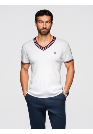 Uomo in piedi con le mani nelle tasche che indossa un maglione bianco a coste a scollo a V con maniche corte, righe color borgogna e pantaloni blu navy su sfondo neutro.