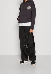 Osoba oblečená v tmavé mikině s bílým logem, širokých černých džínách, černobílé tenisky, držící černou tašku přes rameno s nastavitelným popruhem.