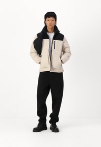 Beige gepolsterte Jacke mit schwarzen Akzenten und Kapuze, ausgestattet mit mehreren Taschen und einem Reißverschluss. Kombiniert mit lockeren schwarzen Hosen und schwarzen Stiefeln.