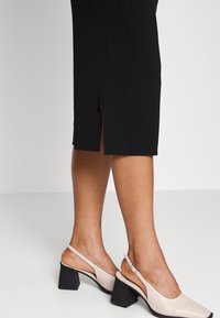 Beige slingback-korkokengät neliömäisellä varpaalla, joissa on paksu musta korko. Yhdistetty mustaan polvipituiseen hameeseen, jossa on sivuaukko.