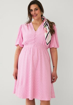 Kvinde iført en pink kjole med hulmønster, puffede ærmer og stofbetrukne knapper, accessoriseret med et blomsterprintet tørklæde i håret.