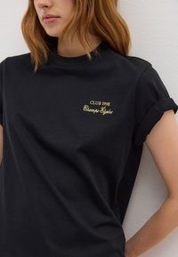Camiseta de algodón negra con mangas enrolladas y bordado dorado que dice "CLUB 1998 Champs-Élysées". Textura suave y corte relajado.