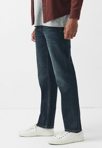 Jeans de denim azul oscuro con un corte recto, combinados con zapatillas blancas. La textura es de denim, contrastando con una camiseta gris y una capa de camisa burdeos.