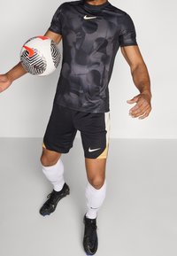 Černé sportovní tričko s krátkým rukávem a jemným abstraktním vzorem, spárované s černými šortkami s zlatými akcenty a bílým logem Nike. Fotbalový míč v ruce.
