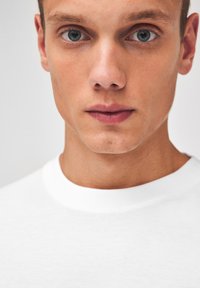 T-shirt blanc à col rond, gros plan sur le visage d'un modèle masculin, présentant une peau claire, des yeux bleus et des cheveux courts sans motifs ou accents distincts.