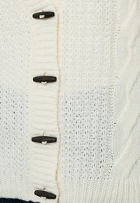 Närbild av vitt stickat tyg med ribbstruktur, kabelstickningsmönster och fyra mörkbruna togglar sydda med vit tråd.