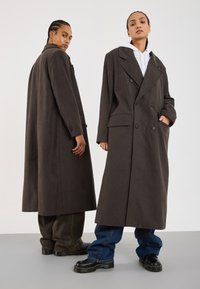 Brown double-breasted overcoat with large lapels, side pockets, and loose fit, paired with dark jeans and chunky black boots.