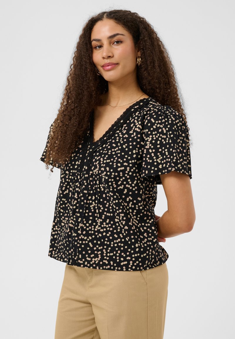 Femme aux longs cheveux bouclés portant un chemisier noir à manches courtes avec des taches beiges et un pantalon beige, debout, les mains dans le dos.