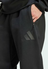 Black jogger pants made of soft fabric, featuring a zippered side pocket and a subtle, glossy three-stripe logo on the thigh.