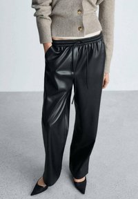 Pantalon en simili cuir noir à coupe décontractée, avec ceinture élastique et cordon de serrage, associé à un pull boutonné gris clair côtelé.