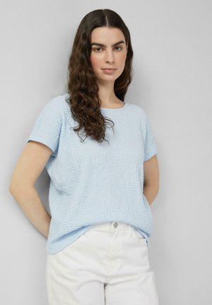 Jeune femme aux longs cheveux bouclés, debout les mains derrière le dos, portant un haut à manches courtes bleu clair texturé et un pantalon blanc.