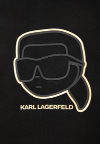 Stylizowany zarys twarzy w dużych okularach przeciwsłonecznych i z kucykiem, nad napisem "KARL LAGERFELD" na czarnym tle.