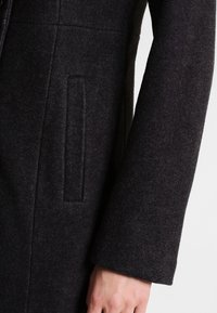 Primo piano di una manica e tasca di un cappotto di lana grigio scuro con una mano parzialmente visibile in basso a destra.